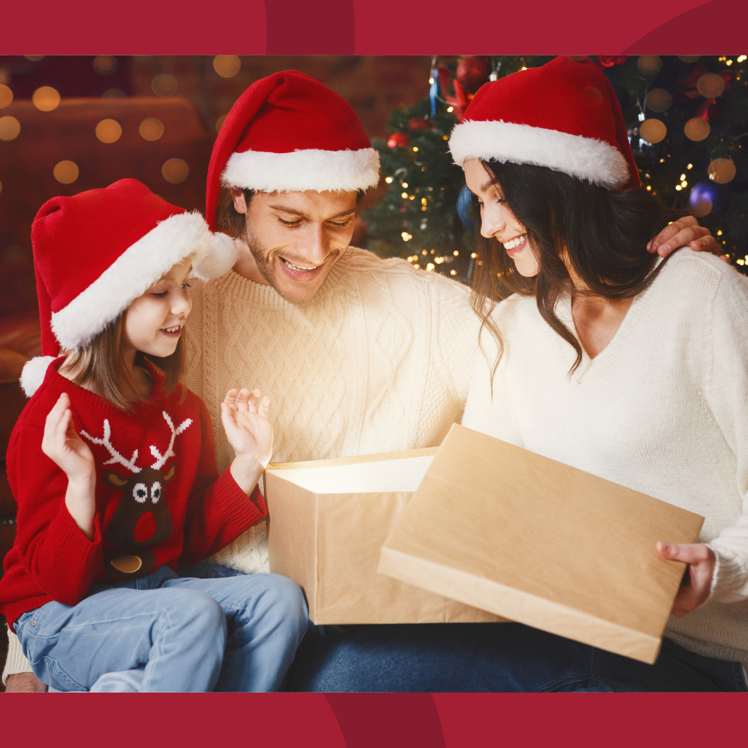 A young family is sitting in front of Christmas tree while wearing Santa hats and looking in a glowing gift box.